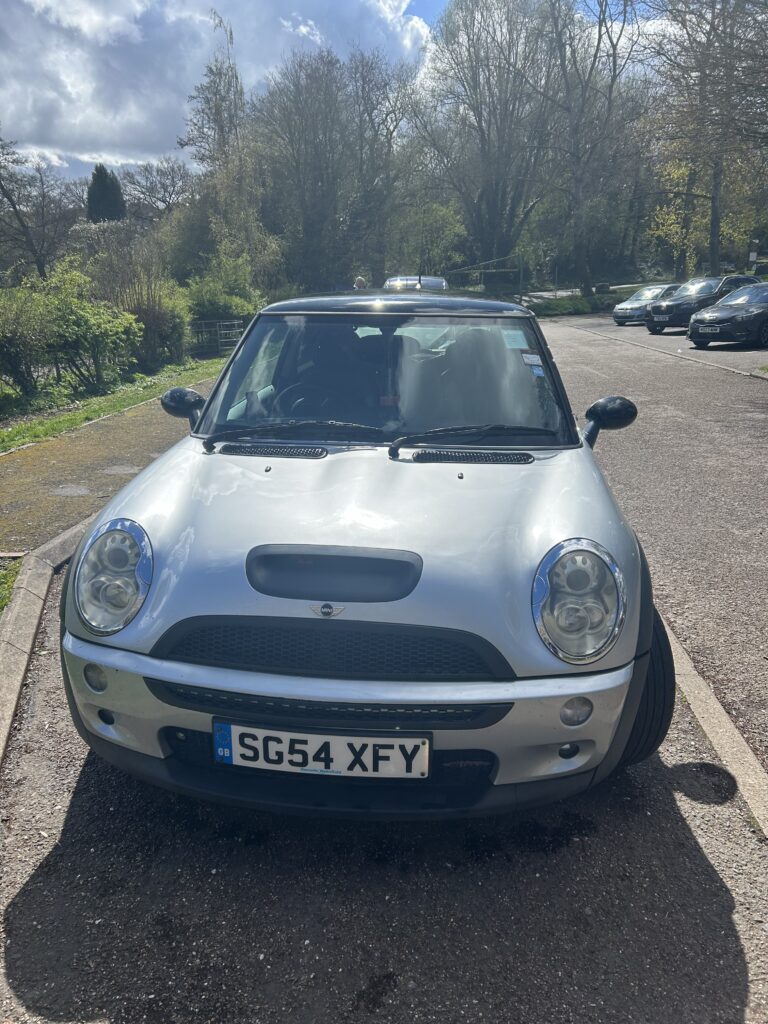 A silver Mini Cooper from a mini trader is parked at an angle in a sunny outdoor lot, front wheels turned right. The car’s UK license plate reads “SG54 XFY.” Trees and other parked cars are visible under a partly cloudy sky.