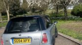 A silver Mini car with a black roof is parked beside a curb under a blossoming tree. The car, featured on Mini Trader, has a UK license plate “SG54 XFY.” Green grass, a wooden fence, and more trees appear in the sunny background.