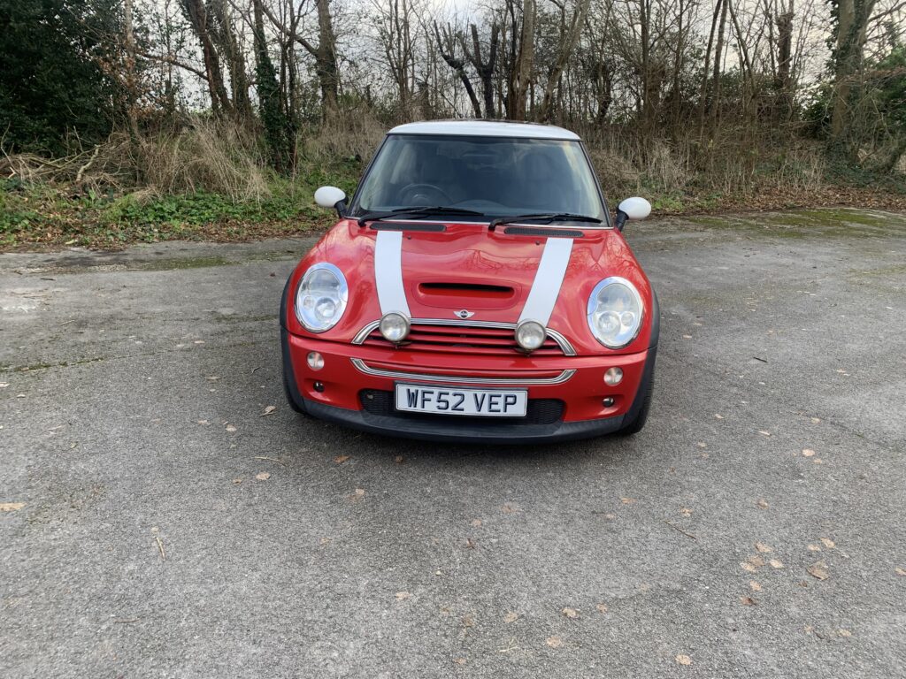 A red Mini Cooper mini car with white racing stripes and extra spotlights on the hood is parked on a concrete surface, surrounded by leafless trees and overgrown grass. The license plate reads "WF52 VEP." The weather appears cloudy and cool.