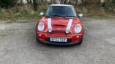 A red Mini Cooper mini car with white racing stripes and extra spotlights on the hood is parked on a concrete surface, surrounded by leafless trees and overgrown grass. The license plate reads "WF52 VEP." The weather appears cloudy and cool.
