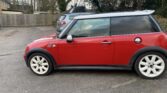 A red Mini Cooper with a white roof and white wheels, listed on Mini Trader, is parked on a paved surface. Another mini car and some houses are visible in the background amid trees and a wooden fence beneath an overcast sky.