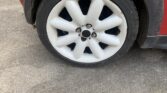 Close-up of a mini car’s front wheel with a white rim featuring eight rounded, thick spokes, mounted with a black tire. The red car body is partially visible, parked on rough, light gray pavement.
