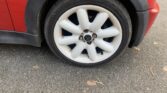Close-up of a white alloy car wheel with visible rust spots around the bolts, attached to a red mini car. The tire rests on grey asphalt with scattered dry leaves, and the mini’s black fender and part of its red body are also in view.