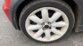 A close-up of a mini car’s front wheel with a white, ten-spoke alloy rim. The tire is slightly worn with dirt around the rim and wheel well. The red and black mini body is partly visible above, parked on a gray asphalt surface.