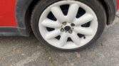 Close-up of a white alloy wheel on a red mini car, showing eight large rounded spokes. The black tire has visible tread and some dirt on the rim. The mini is parked on gray asphalt with small bits of debris nearby.
