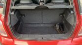 The image shows the open trunk of a red mini hatchback car. The trunk is empty, except for a metal tool on the right side. The rear seats and car interior, including the dashboard and steering wheel, are visible through the open trunk.