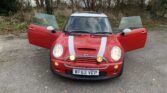 A red mini car with white racing stripes is parked on grey pavement with both front doors open. The headlights and fog lights shine brightly. Trees and bushes are in the background, and the license plate reads "WF52 VEP.