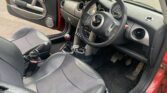The image shows the interior of a compact Mini car with black and gray seats, a black leather steering wheel, manual gear shift, round air vents, and a large central speedometer on the dashboard. The driver’s door is open and the floor is carpeted.
