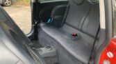 The image shows the back seat of a small red mini car with dark gray upholstery. The seat has two seatbelt buckles in the center, a cup holder on the floor between the front seats, and part of the front passenger seat. Bushes are visible outside.