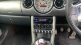 The image shows the center console of a MINI car interior. The speedometer is centrally located above round air vents, with a stereo below displaying "CHECKING." Below are control knobs and toggle switches. The gear shift, with a Union Jack design, is in the foreground.
