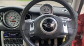 View from the driver’s seat of a mini car, showing the black steering wheel with a Mini logo, large central speedometer, smaller gauges, dashboard controls, and exposed foot pedals. Part of the red door and exterior street are visible.
