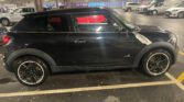 A black Mini Paceman car is parked in an indoor parking lot. This sporty Mini car features two doors, alloy wheels, and a polished surface with visible reflections. Other vehicles and shops are seen in the background under fluorescent lights.
