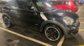 A black Mini Cooper with silver trim is parked in an indoor parking lot. The mini car features black alloy wheels and a visible "ALL4" badge on the door. In the background, a red car sits across yellow-lined parking spaces.