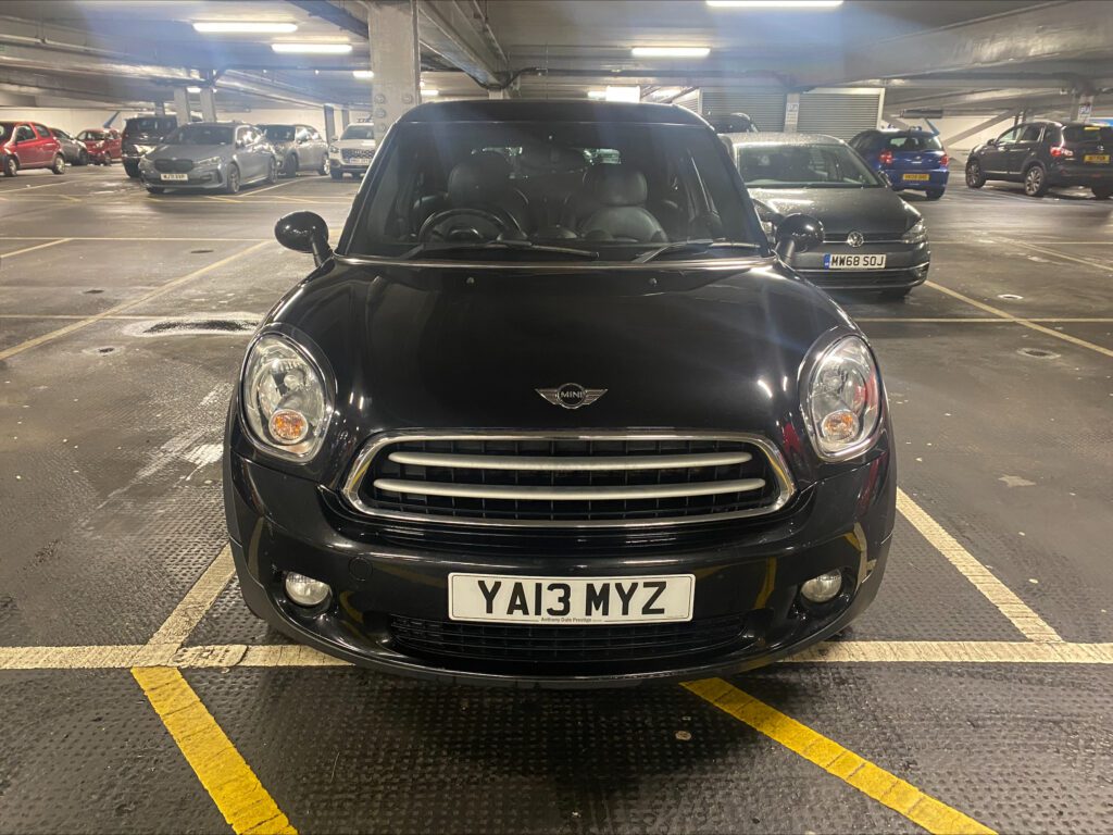 A black mini car is parked head-in inside a multi-story car park. The mini faces the camera, displaying a "YA13 MYZ" plate. Other cars are seen in the background under artificial lighting, with concrete pillars and marked spaces.