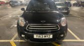 A black mini car is parked head-in inside a multi-story car park. The mini faces the camera, displaying a "YA13 MYZ" plate. Other cars are seen in the background under artificial lighting, with concrete pillars and marked spaces.