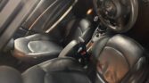 A photo shows the black leather interior of a mini car, featuring the driver and front passenger seats, steering wheel with a MINI logo, gear shift, handbrake, cupholders, and door panel with silver trim. The cabin is illuminated by soft lighting.