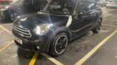 A sleek black Mini Cooper, a standout mini car with tinted windows and black alloy wheels, is parked at an angle in an indoor garage. Its headlights are on, and other cars can be seen in the background.