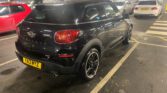 A black MINI Paceman mini car is parked in a multi-story car park with other vehicles nearby. The car has a yellow license plate "YA13 MYZ," distinctive rear lights, chrome accents, and is positioned within white lined, numbered spaces.