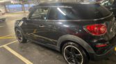 A shiny black Mini Cooper Paceman, listed on Mini Trader, is parked in a multi-level garage. The mini car features black alloy wheels, red tail lights, and silver door handles. Garage lights reflect off its surface as other vehicles park nearby.