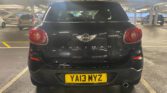 A sleek black mini car, the Mini Paceman Cooper, is parked in an indoor garage. Viewed from behind, its yellow "YA13 MYZ" plate stands out among other cars and concrete pillars under fluorescent lighting.