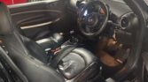 The image shows the black leather interior of a Mini, viewed from the driver’s side door. Features include a round central speedometer, manual gear stick, key in the ignition, and a red car outside—perfect for any Mini trader.