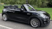 A sleek black Mini Cooper convertible with the top down is parked on a paved road. Lush green trees and grass surround the mini car under a partly cloudy sky. The phone screen displays the time: 3:29 PM, 18/06/2024.