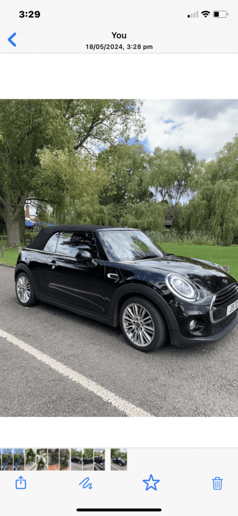 A black Mini Cooper convertible, featured for mini trader enthusiasts, is parked on a road beside a grassy area with trees under a cloudy sky. The car’s top is up, highlighting its headlights and alloy wheels. The time and date appear at the top of the image.