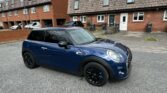 A blue mini car with black wheels is parked on a residential street lined with brick and tiled terraced houses. The sky is mostly cloudy, and the empty street highlights this stylish Mini, perfect for any mini trader enthusiast.