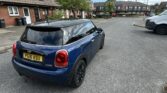 A blue mini car with a black roof and UK license plate "PG16 ASU" is parked on a residential street beside brick houses with tiled roofs. The sky is partly cloudy, and another vehicle is ahead. Yellow road markings are visible.