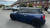 A blue mini car with black wheels is parked on a residential street under a partly cloudy sky. Row houses, a white van, and other cars are visible in this urban scene with greenery nearby. Perfect for any mini trader or enthusiast.