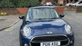 A dark blue Mini car with a black roof is parked on a residential street facing forward. The license plate reads "PG16 ASU." Red-brick houses and parked cars are visible in the background, along with trees and a cloudy sky.