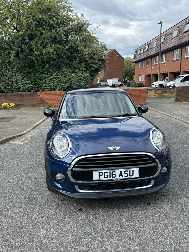 A dark blue Mini car with a black roof is parked on a residential street facing forward. The license plate reads "PG16 ASU." Red-brick houses and parked cars are visible in the background, along with trees and a cloudy sky.