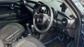 The image shows the clean interior of a mini car with the driver’s door open. It has black fabric seats, a manual gear stick, and a dashboard with circular air vents. The steering wheel features a MINI logo, and blue-trimmed floor mats are visible.
