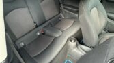 View of a mini car’s back seat area with black fabric upholstery, three seatbelt buckles, headrests, a cup holder on the floor, and blue-edged floor mats. The front passenger seat is visible; outside, a sidewalk and another car are seen through the window.