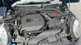 A photo shows the engine bay of a blue MINI car with its hood open. The engine cover, labeled “MINI” and featuring the MINI logo, sits among visible parts like the air filter box and coolant cap. Ideal for any MINI trader, the car is parked on a paved surface.