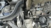 Close-up view of a mini car engine bay, showing various metal and plastic components, cables, hoses, and connectors. The area appears dusty and slightly dirty, with complex mechanical parts and some exposed inner bodywork. No people are visible.