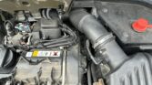A close-up view of a mini car engine with the hood open, showing various components including hoses, wiring, and the air intake system. The engine bay appears somewhat dusty, and a red cap—likely for the battery or coolant—is visible on the right.