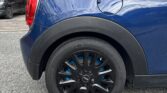 A close-up of the rear left side of a blue mini car parked on a street. The focus is on the black alloy wheel with blue brake calipers. Part of the taillight and rear window are visible, with houses and another parked car in the blurred background.