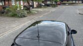 A blue mini car with a reflective sunroof is parked on a quiet residential street. Tree branches and clouds are mirrored on the roof as rows of brick houses with gardens and greenery line both sides under a partly cloudy sky.
