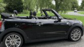 A sleek black Mini Cooper convertible with its roof down is parked roadside. This stylish mini car sits against green grass and leafy trees beneath a cloudy sky, with open windows and both front seats visible.