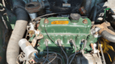 Close-up view of a vintage mini car engine with a green valve cover, silver ignition coil, black spark plug wires, a large flexible hose, and various metal parts. Some areas appear rusted and weathered, showing this mini trader’s years of use.