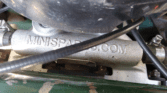 Close-up of a metallic mini car part with "MINISPARES.COM" embossed on it. Black cables and clamps are attached on both sides, with a black rubber component above and a chipped green painted surface in the background.