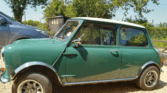 A classic green Mini car with a white roof is parked on a gravel driveway. Trees and a wooden shed are visible in the background, while part of a gray car sits nearby. The sun is shining, casting clear shadows. Perfect for any Mini trader or enthusiast.