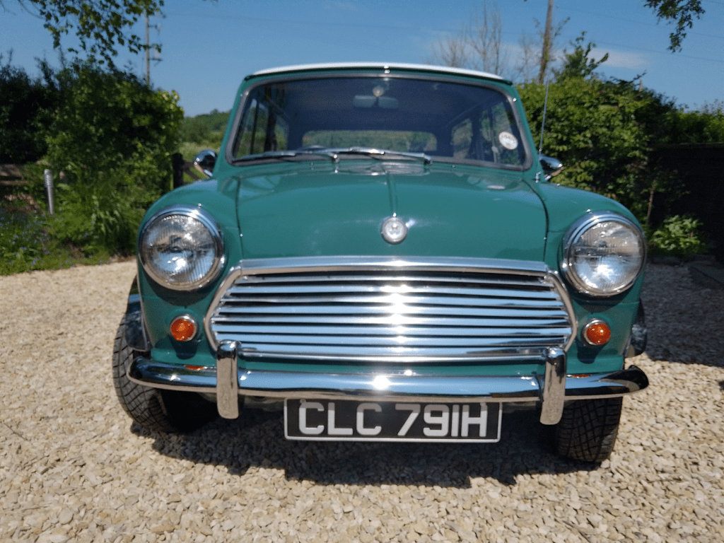 A classic green mini car is parked on a gravel driveway, facing forward. The car has chrome bumpers, round headlights, and a black license plate reading "CLC 791H." Lush greenery and a clear blue sky are visible in the background—perfect for any Mini trader.