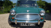 A classic green mini car is parked on a gravel driveway, facing forward. The car has chrome bumpers, round headlights, and a black license plate reading "CLC 791H." Lush greenery and a clear blue sky are visible in the background—perfect for any Mini trader.