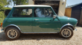 A classic green mini car with a white roof is parked on a gravel driveway. The car has chrome accents, round headlights, and alloy wheels. Behind it is a small garage with an open door and a wooden shed to the right. Trees and bushes border the scene.