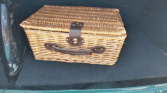 A rectangular wicker picnic basket with a brown leather strap and handle sits on a dark carpeted surface, likely in the trunk of a mini car. The basket's lid is secured, and the car’s green metal frame is partly visible at the bottom.
