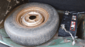 A mini car trunk with a dirty, worn spare tire lies flat beside a black car battery with exposed terminals and wires. The green metal floor shows rust and wear, while dark carpeting is partially visible in the upper part of the trunk.
