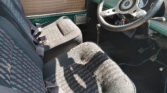 Interior of a classic mini car showing two front seats with patterned upholstery, seat belts, a simple dashboard with a large black steering wheel, manual gear stick, and brown door panel. Sunlight casts shadows across the seats.