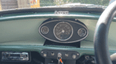 Close-up of a vintage mini car dashboard viewed from the driver's seat. The central speedometer reads 28898 miles, flanked by smaller gauges. Keys hang from the ignition, and sunlight brightens the retro green and black interior.